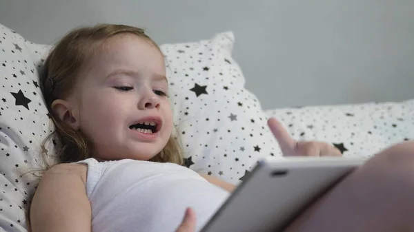 Маленький счастливый малыш играет на планшете, лежа на кровати. Ребенок смотрит на канал детей через сенсорный монитор и смеется. Обучение дошкольника дистанционно в режиме онлайн — стоковое фото