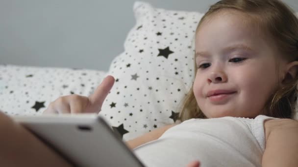A kis boldog baba tablettázik, miközben fekszik az ágyon. A kölyök nézi a gyerekek csatornáját az érintőképernyőn keresztül és nevet. Tanítani egy óvodást távolról online — Stock videók