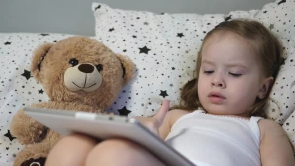 Das kleine glückliche Kind spielt mit einem Tablet, während es auf einem Bett mit einem Teddybär liegt. Das Kind beobachtet den Kinderkanal über den Touchmonitor. Unterricht eines Vorschulkindes aus der Ferne online. Nahaufnahme — Stockvideo