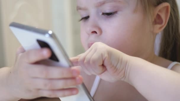 A kislány okostelefonon játszik. A gyermek távolról tanul a telefonos alkalmazásban. Óvodás az interneten — Stock videók