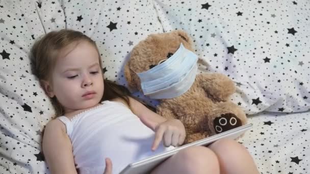 Ein kleines Kind mit einem Bären in Maske spielt auf einem Tablet, während es auf dem Bett liegt. Kovid-19. Kind wird zu Hause unter Quarantäne gestellt. Fernunterricht für Vorschulkinder. Moderne Technologien — Stockvideo