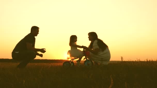 Egy gyerek sziluettje biciklin a szüleivel naplementekor. Anya, apa és a baba játszanak az esti parkban. Boldog családi életet. Csapatmunka — Stock videók