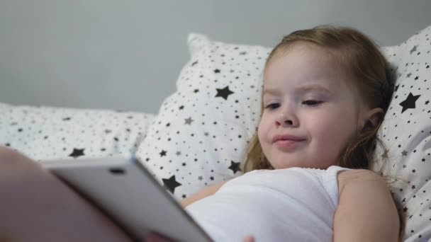 Kleine gelukkige baby speelt tablet terwijl hij op het bed ligt. Kid kijkt naar de kinderen kanaal door de touch monitor en lacht. Leren van een kleuter op afstand online — Stockvideo