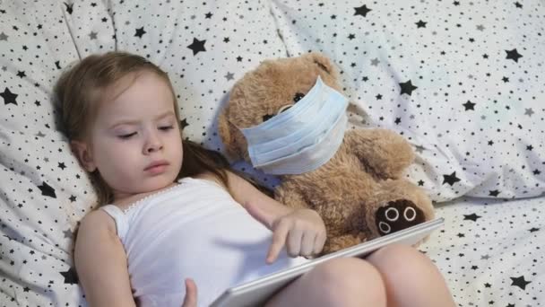 Uma criança pequena com um urso em uma máscara joga um tablet enquanto está deitada na cama. Kovid-19. O miúdo está em quarentena em casa. Aprendizagem à distância para pré-escolares. Tecnologias modernas — Vídeo de Stock