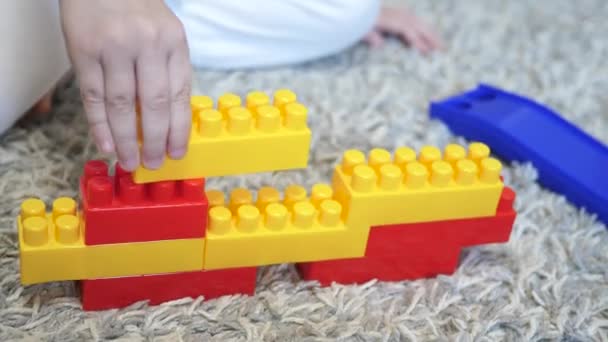 Un bambino gioca nel costruttore seduto sul pavimento. Bambina giocattolo. Sviluppo e pensiero dell'apprendimento prescolare. Un bambino piccolo costruisce da mattoni multicolori. Primo piano — Video Stock