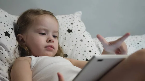 Маленький счастливый малыш играет на планшете, лежа на кровати. Парень смотрит канал для детей через сенсорный монитор и смеется. Обучение дошкольника дистанционно в режиме онлайн — стоковое фото