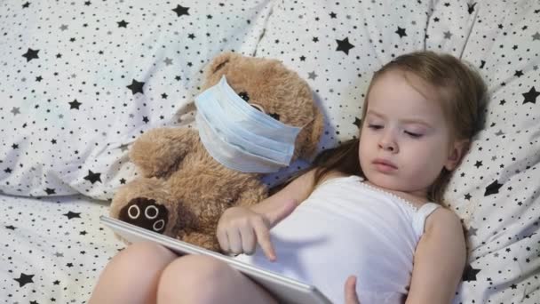 Ένα μικρό παιδί με μια αρκούδα σε μια μάσκα παίζει ένα δισκίο, ενώ βρίσκεται στο κρεβάτι. Κόβιντ-19. Το παιδί είναι σε καραντίνα στο σπίτι. εξ αποστάσεως εκπαίδευση για παιδιά προσχολικής ηλικίας. Νέες τεχνολογίες — Αρχείο Βίντεο