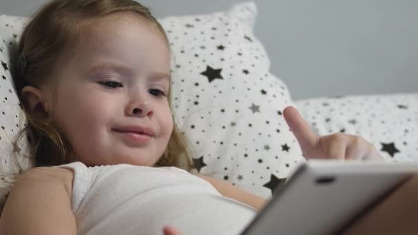 A kis boldog baba tablettázik, miközben fekszik az ágyon. A kölyök nézi a gyerekek csatornáját az érintőképernyőn keresztül és nevet. Tanítani egy óvodást távolról online — Stock videók
