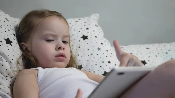 Küçük mutlu bebek yatakta yatarken tablet oynuyor. Çocuk dokunmatik monitörden çocukların kanalını izliyor ve gülüyor. Bir anaokulu öğrencisine uzaktan eğitim vermek. — Stok fotoğraf