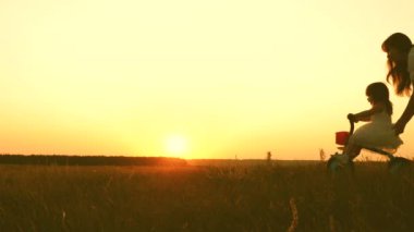 Anne küçük bir çocuğa gün batımında parkta bisiklet sürmeyi öğretiyor. Takım çalışması. Siluet aile yürüyüşü