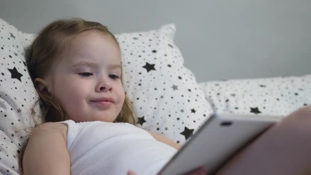 Pequeno bebê feliz está jogando tablet enquanto deitado na cama. Kid observa o canal das crianças através do monitor de toque e ri. Ensinar um pré-escolar remotamente online — Vídeo de Stock