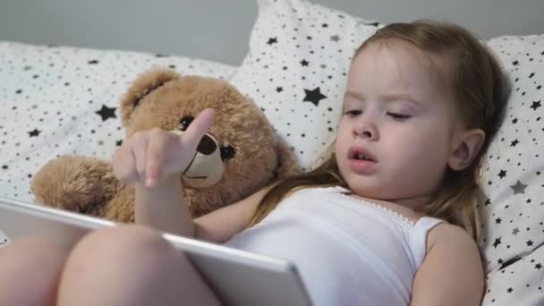 A criança feliz está a jogar um tablet. Uma menina jaz na cama com um ursinho de pelúcia — Vídeo de Stock