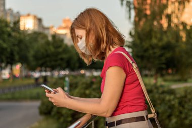 Açık havada elinde akıllı bir telefonla tıbbi koruyucu maskeli genç bir kadın. 2020 Coronavirus salgınında karantina.