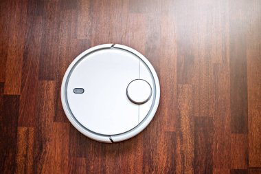 Beyaz bir robot süpürge kahverengi bir iç mekanda yerleri temizliyor.