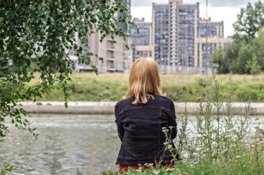 Genç bir kadın sırtı nehir kıyısında, şehir manzaralı oturuyor..