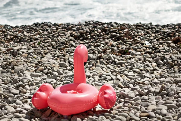 Inflatable toy pink flamingo lies on a pebble beach against the background of the sea. Holiday concept by the sea.