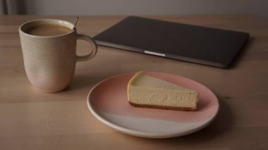 Kahvaltı tabağında nefis bir cheesecake. Tahta bir masada. Yanında da laptopun yanında bir fincan kahve.