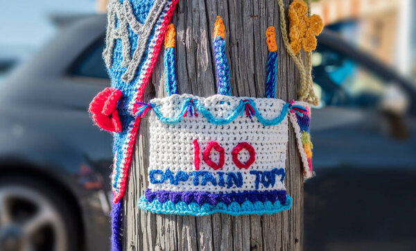 Crochet birthday cake for Captain Tom celebrating his 100th birthday put on a street post with a car in a residential area in the background