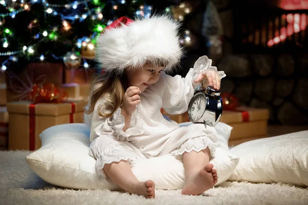 サンタ クロースを待っている暖炉のクリスマス ツリーの下に枕に目覚まし時計を持つ少女 — ストック写真