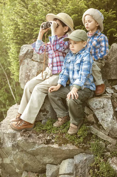 Niños viajeros mirando a la distancia a través de prismáticos, sentados en una gran roca —  Fotos de Stock