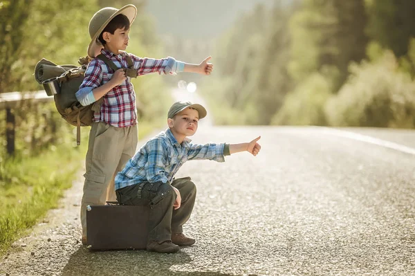 Niños viajeros en una carretera de montaña con una mochila y una maleta —  Fotos de Stock