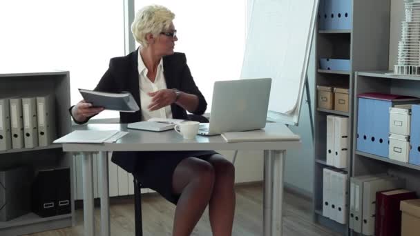 在办公室工作的女人 那个女人 可能是头 在办公室的工作室里 在她的办公桌上 我们看到笔记本电脑 报告文件夹 她在文件中记笔记 检查电子邮件 她穿着西装 — 图库视频影像