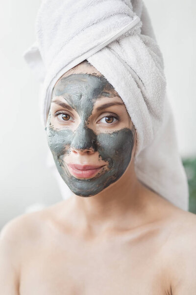 Face Skin care. Attractive Young Woman Wrapped in Bath Towel, applying clay mud mask to face. Skin care concept. Girl taking care of complexion. Beauty treatments