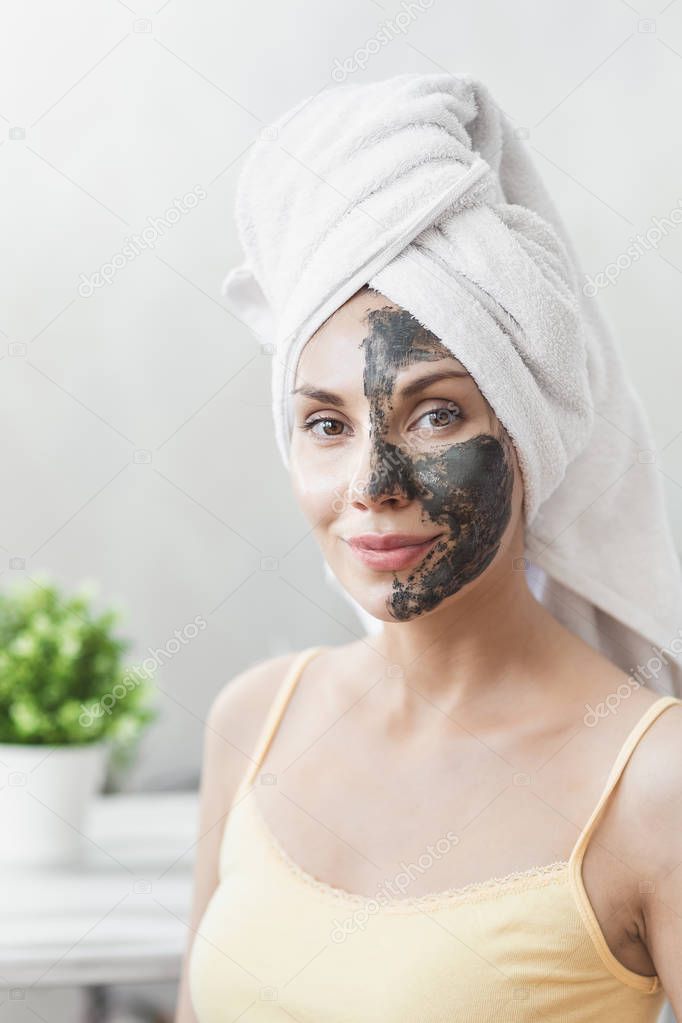 Face Skin care. Attractive Young Woman Wrapped in Bath Towel, applying clay mud mask to face. Skin care concept. Girl taking care of complexion. Beauty treatments