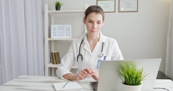 Médico falando com o paciente usando aplicativo de computador de bate-papo virtual. Terapeuta médica feminina usa vestido médico, fone de ouvido vídeo chamando paciente distante no laptop. Conceito de serviços de saúde remotos de telemedicina — Vídeo de Stock