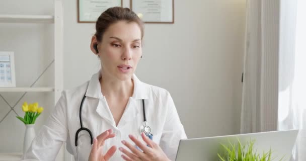 医生使用虚拟聊天电脑应用与病人交谈。女医生穿着医疗服，带着耳机，用手提电脑给远方的病人打电话。远程医疗远程保健服务概念 — 图库视频影像