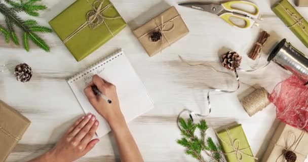 Vista superior, la cámara se mueve lentamente sobre la mesa de madera, en la que las decoraciones de Navidad, regalos, árbol de Navidad, niña escribe en los planes de mesa de Año Nuevo para el nuevo 2021. Navidad y año nuevo concepto — Vídeo de stock