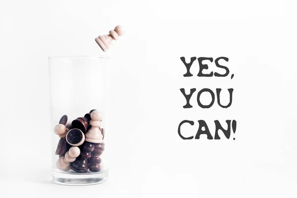 Motivasyon alıntısı. Evet, yapabilirsin. Beyaz arka planda satranç taşlarıyla dolu bir cam bardak üzerinde. Beyaz piyon camdan atlıyor..