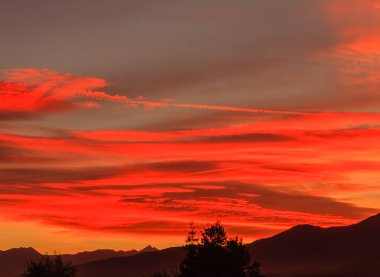 Sıcak bir günbatımı, kırmızı bir bulut ve bir yoğunlaşma izi. Gün batımında dağların ve ağaçların çizdiği bir manzara. 