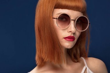 Portrait of beautiful redhead young woman wearing fashionable sunglasses. Girl in glamour makeup. Haircut with fringe. Blue background. clipart