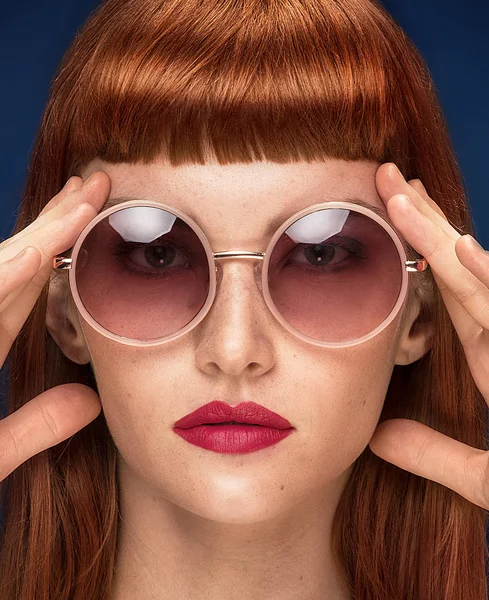 Güzel kızıl saçlı Genç kadın moda güneş gözlüğü takıyor portresi. Glamour makyaj kız. Saçak ile saç kesimi. Mavi arka plan.