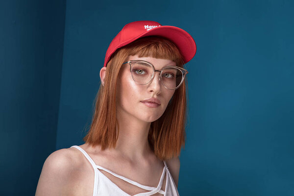 Portrait of attractive young redhead woman with fashionable eyeglasses and cap. Girl looking at camera. Blue background.