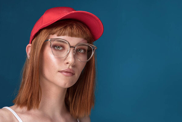 Portrait of attractive young redhead woman with fashionable eyeglasses and cap. Girl looking at camera. Blue background.