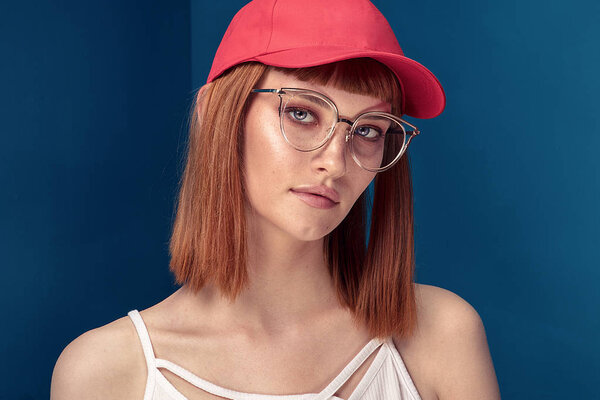 Portrait of attractive young redhead woman with fashionable eyeglasses and cap. Girl looking at camera. Blue background.