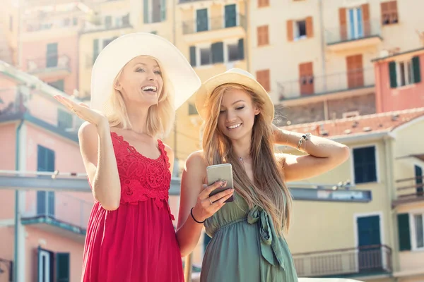 Happy female wears summer hat and fashionable dress spends recreation time with best friend, walk on italian city streets,using smart phone. Summer sunny day.