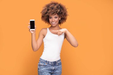 Hareket eden telefon tutarak ve sarı arka plan üzerinde duran boş ekranda gösterilen gülümseyen güzel bir Afrikalı kadınla afro saç modeli, fotoğrafı.
