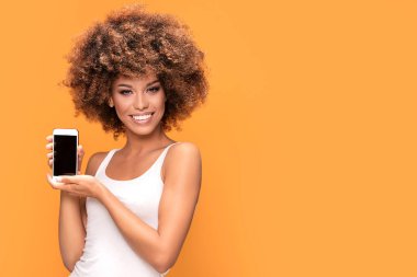 Cep telefonu ile boş perde, sarı arka plan üzerinde ayakta tutan bir gülümseyen güzel Afrikalı kadın fotoğrafı ile afro saç modeli,.