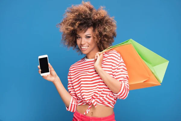 Happy african american woman holding colorful shopping bags and showing mobile phone with empty screen, smiling, posing on blue background.