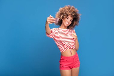 Cep telefonu ve alma selfie, üzerinde mavi arka plan ayakta tutan bir gülümseyen güzel Afrikalı kadın fotoğrafı ile afro saç modeli,.
