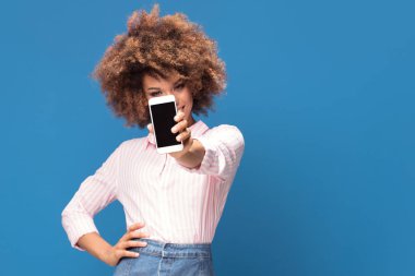 Cep telefonu gösterilen ve gülümseyerek, mavi arka plan üzerinde duran güzel afro bir kadın. Beyaz ve pembe diziler halinde kot mini etek ve zarif forması giyen kız.