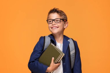 Gülümseyen okul çocuğu turuncu arka plan üzerinde duran, kitaplar, holding sırt çantası ile gözlük.