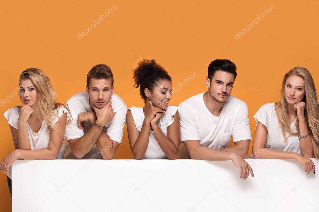 Group of young multi-ethnic beautiful people wearing white shirts, posing together with empty white board. Cheerfull smiling friends.