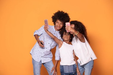 Mutlu renkli üç afro çocuklarda gülümseyerek poz birlikte, gülümseyen, alarak selfie cep telefonuyla elbise.