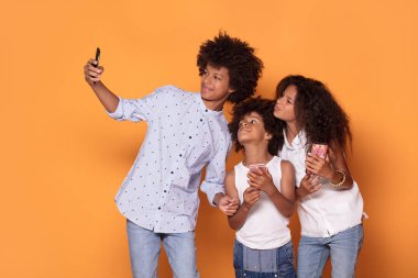 Mutlu renkli üç afro çocuklarda gülümseyerek poz birlikte, gülümseyen, alarak selfie cep telefonuyla elbise.