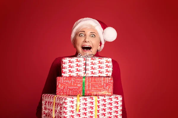 Happy smiling mature woman in red santa claus hat holding stack of gift boxes, on red studio background. Christmas shopping, copy space.