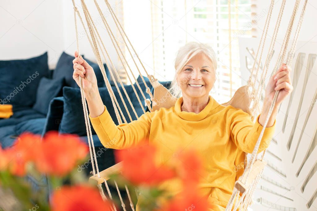 Portrait of beautiful senior woman sitting on a boho swing at home and smiling. Mature lady relaxing in living room. Happy retirement concept.